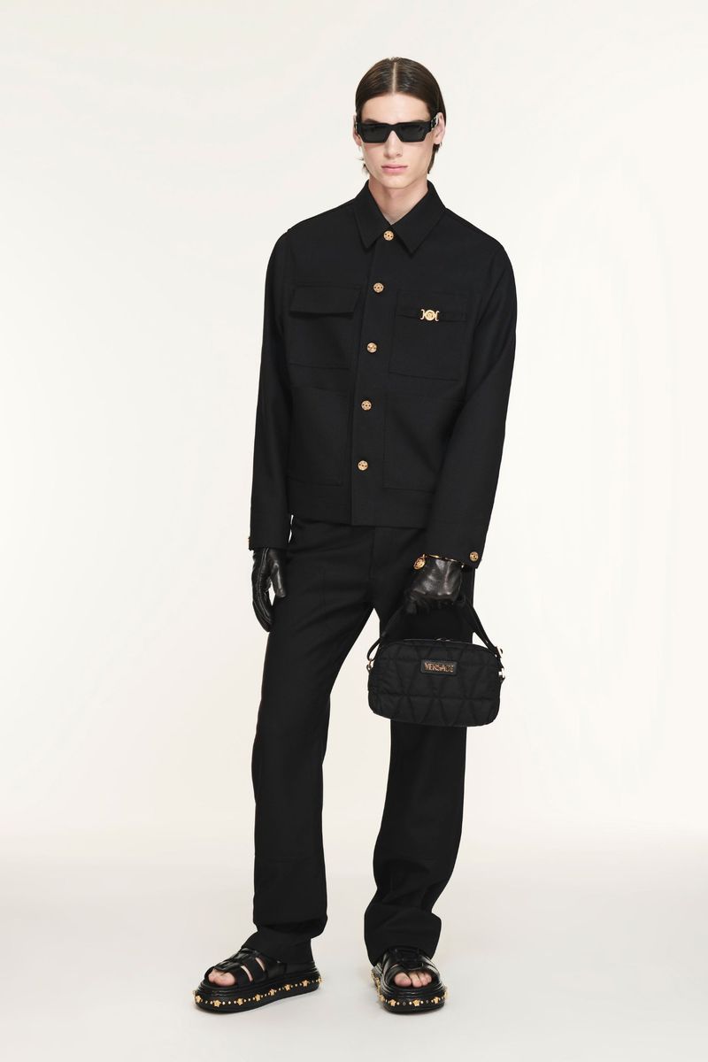 A fair-skinned adult male poses in a black jacket, trousers, sunglasses, and sandals against an off-white backdrop, holding a purse.