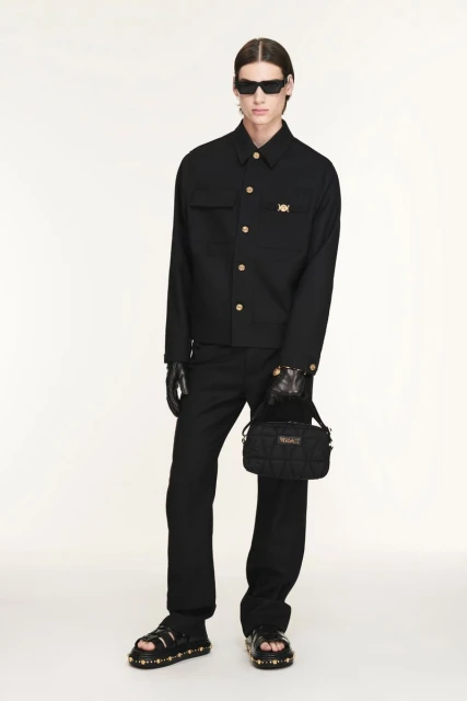 A fair-skinned adult male poses in a black jacket, trousers, sunglasses, and sandals against an off-white backdrop, holding a purse.