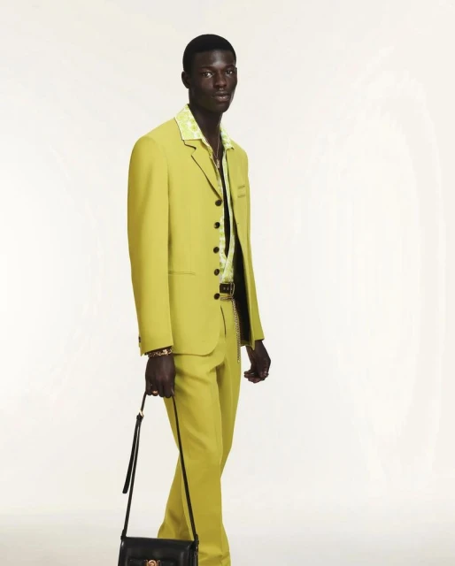 Adult male posing in a chartreuse suit with a black purse, inside a photo studio with bright lighting and a blank white backdrop.