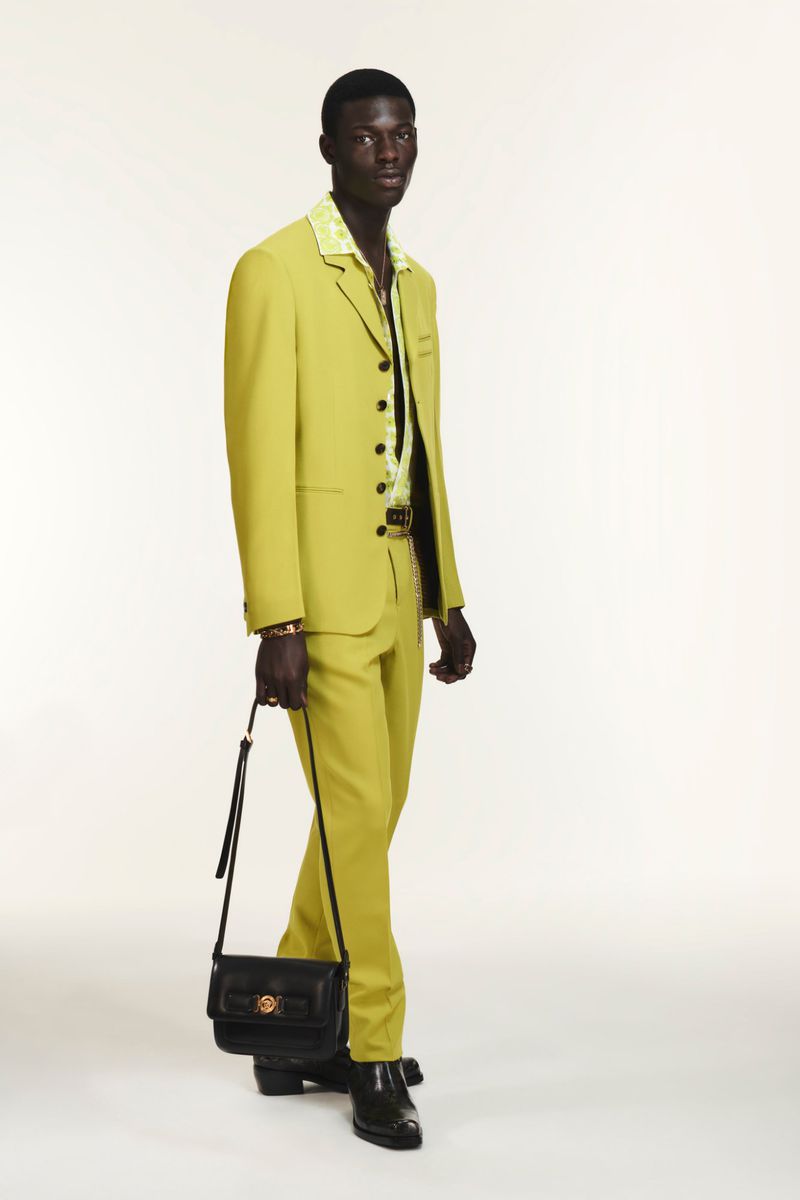 Dark-skinned male model poses in a chartreuse suit with black leather handbag against a clean white backdrop in a studio setting.