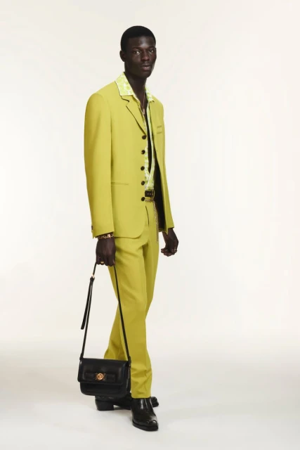 Dark-skinned male model poses in a chartreuse suit with black leather handbag against a clean white backdrop in a studio setting.