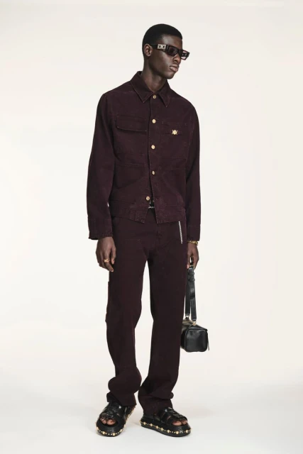 A dark-skinned male fashion model poses in a burgundy corduroy outfit with sandals, sunglasses, and a black bag against a white backdrop.
