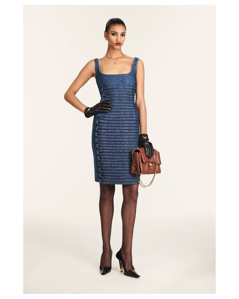 A woman poses in a denim dress with black gloves and heels, holding a brown purse, set against a bright studio background. Fashion.