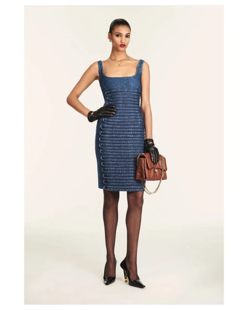 A woman poses in a denim dress with black gloves and heels, holding a brown purse, set against a bright studio background. Fashion.