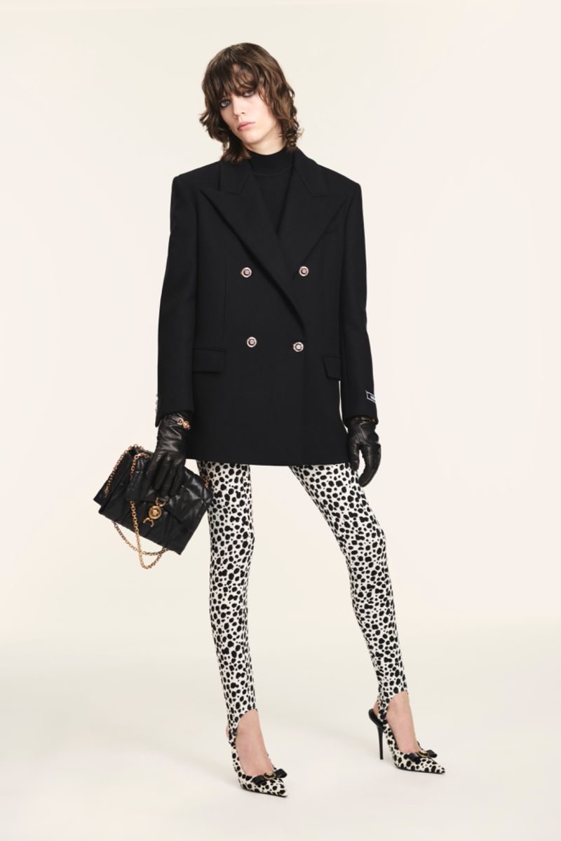 Fashionable woman posing in black blazer and Dalmatian-print leggings with matching high heels and a black quilted shoulder bag on white.