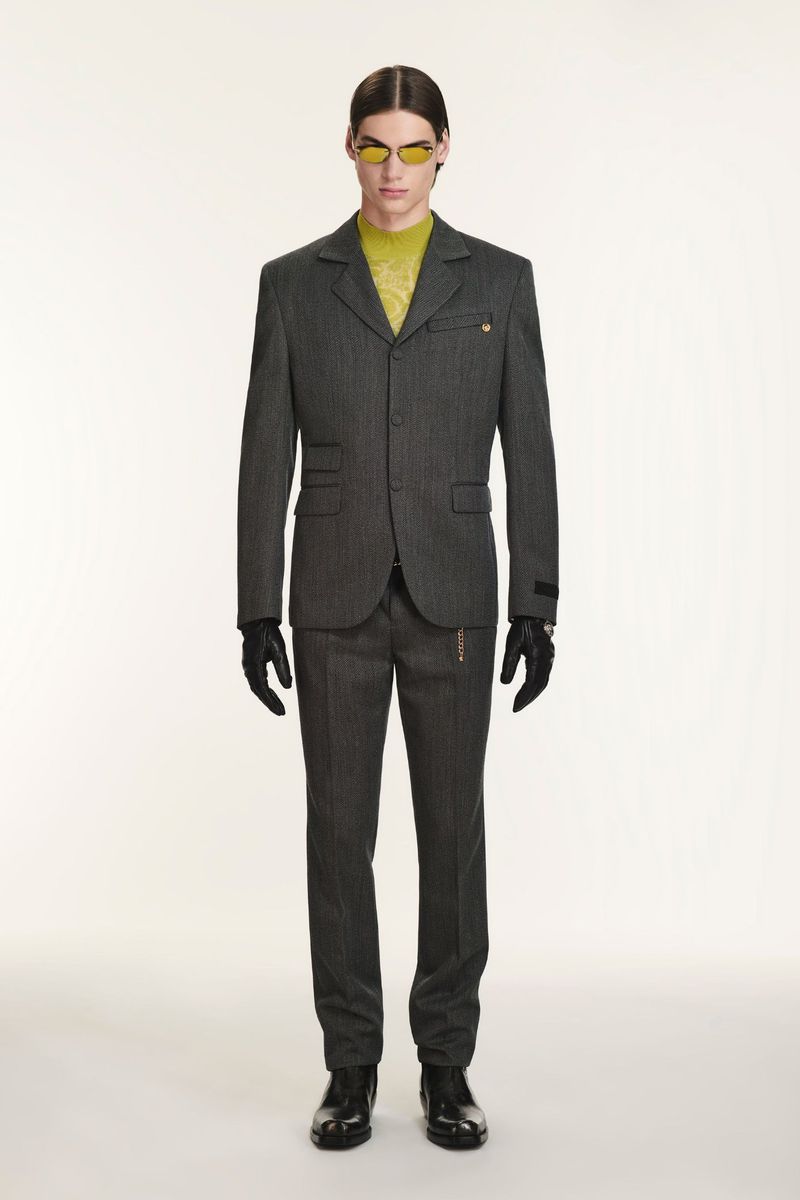 Man in grey suit, yellow top, and sunglasses posing against a white backdrop, showcasing a blend of modern fashion and classic tailoring.