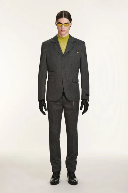 Man in grey suit, yellow top, and sunglasses posing against a white backdrop, showcasing a blend of modern fashion and classic tailoring.