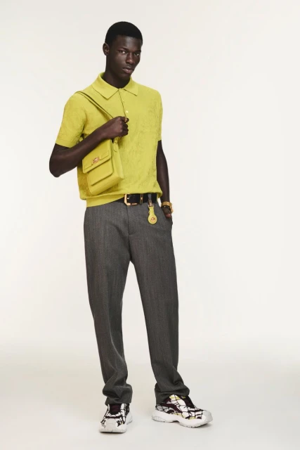 Full-length fashion shot of a Black male model in yellow polo, gray pants, crossbody bag, and sneakers against a plain white backdrop.