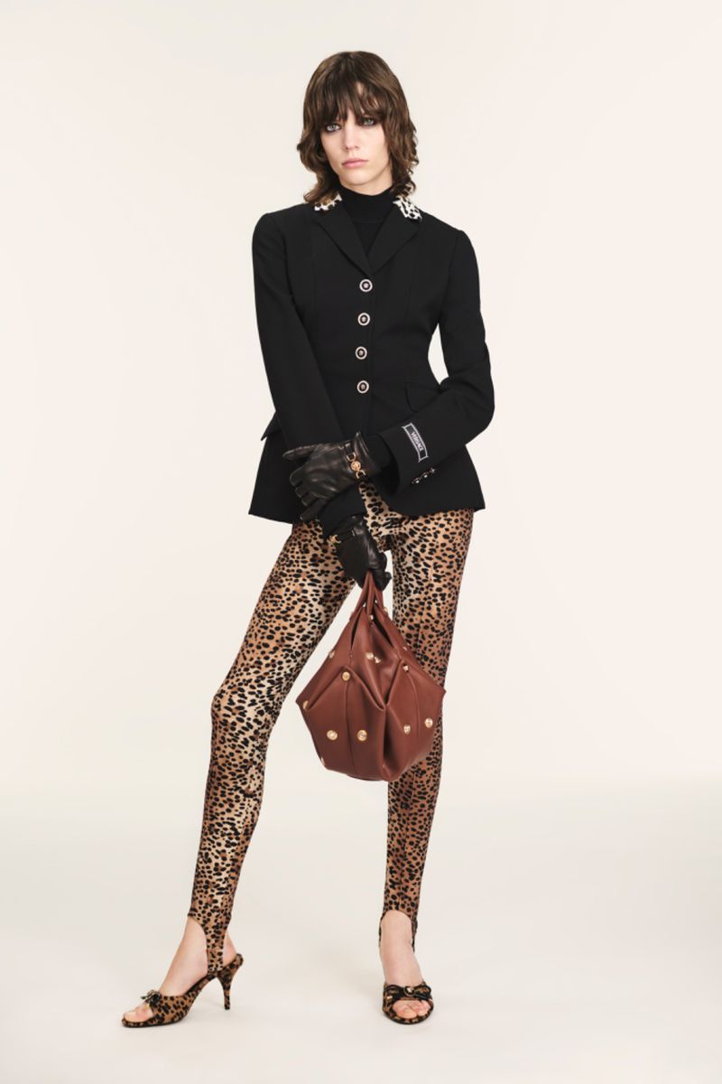 Fashion model posing in black jacket, leopard print leggings and heels, and holding a brown bag against a cream backdrop.
