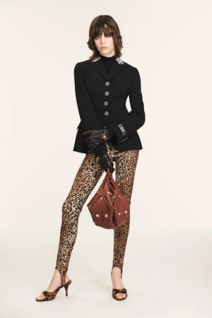 Fashion model posing in black jacket, leopard print leggings and heels, and holding a brown bag against a cream backdrop.