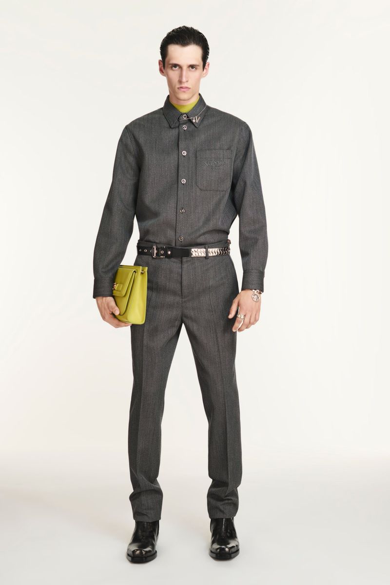Man in grey button-down shirt and trousers, holding lime-green bag, standing in a full shot against a white backdrop in studio lighting.