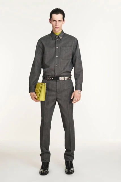 Man in grey button-down shirt and trousers, holding lime-green bag, standing in a full shot against a white backdrop in studio lighting.