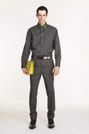 Man in grey button-down shirt and trousers, holding lime-green bag, standing in a full shot against a white backdrop in studio lighting.