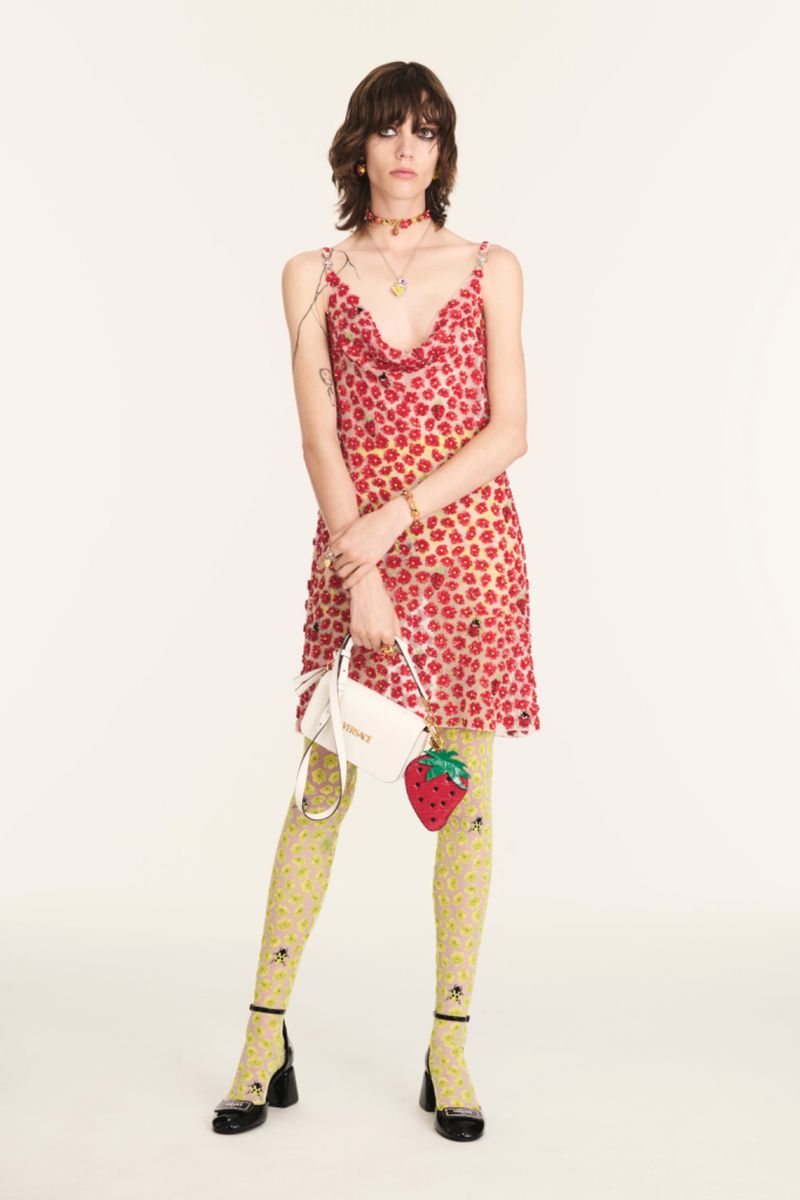 A stylish woman posing in a floral dress with a strawberry purse and patterned tights on a bright background in a studio shot.