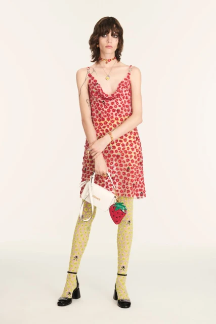 A stylish woman posing in a floral dress with a strawberry purse and patterned tights on a bright background in a studio shot.