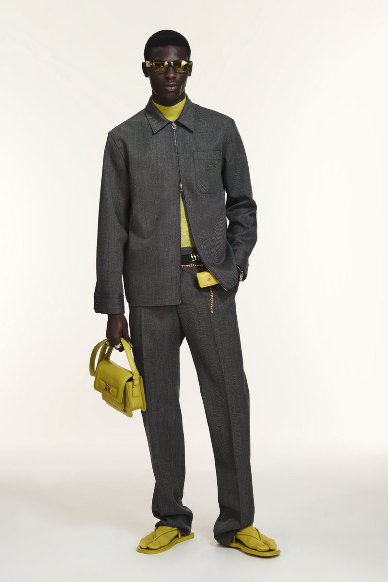 A dark-skinned male fashion model wears a gray pinstripe suit and carries a yellow-green handbag against a plain background.