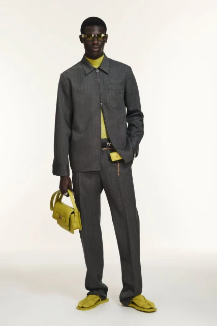 A dark-skinned male fashion model wears a gray pinstripe suit and carries a yellow-green handbag against a plain background.