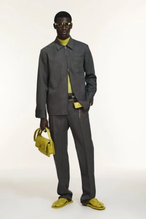 A dark-skinned male fashion model wears a gray pinstripe suit and carries a yellow-green handbag against a plain background.
