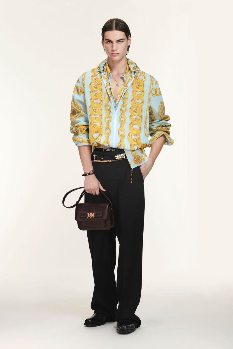 Full-length shot of a young adult male fashion model wearing a patterned shirt, black pants, and carrying a leather handbag in studio.