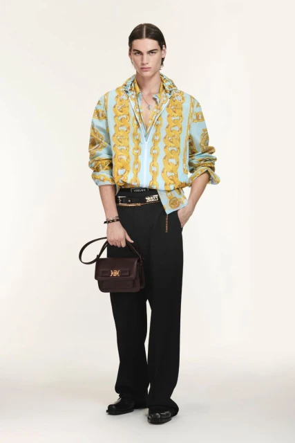 Full-length shot of a young adult male fashion model wearing a patterned shirt, black pants, and carrying a leather handbag in studio.