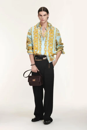 Full-length shot of a young adult male fashion model wearing a patterned shirt, black pants, and carrying a leather handbag in studio.