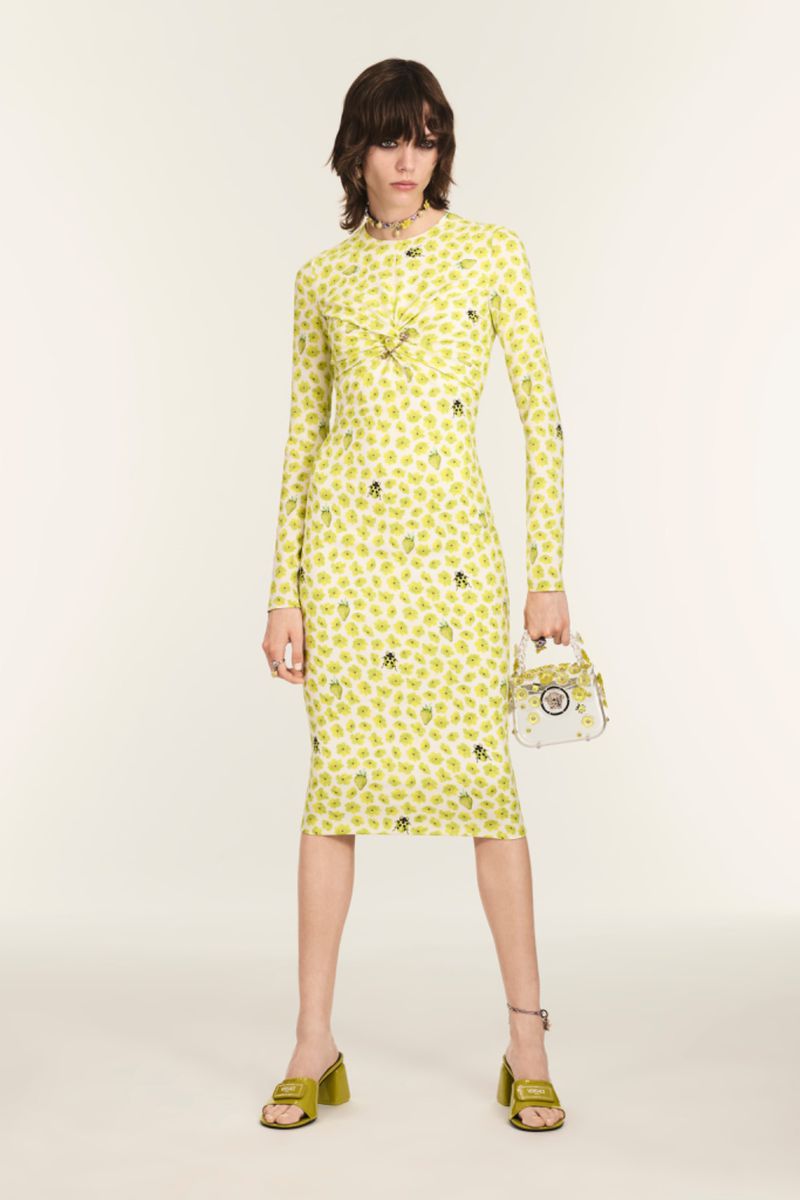 A slender woman poses in a long-sleeved patterned midi dress with olive-green heeled sandals and a clear handbag in a studio setting.