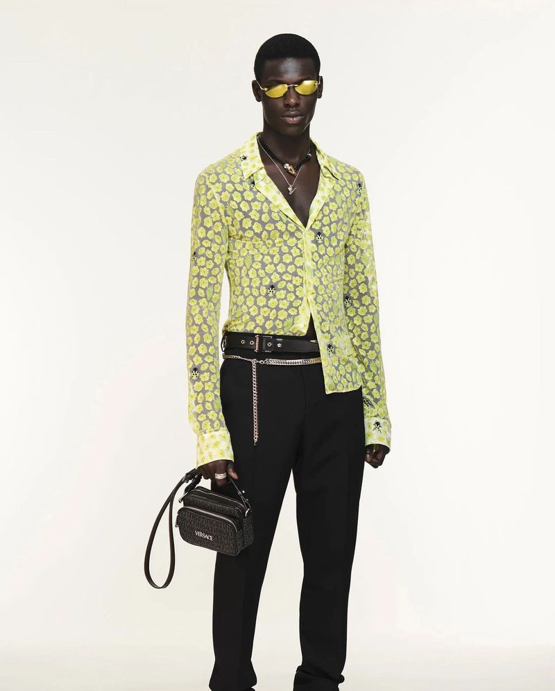 Full-body shot of a Black male model in black pants and a yellow patterned shirt, holding a bag, against a white backdrop in studio lighting.