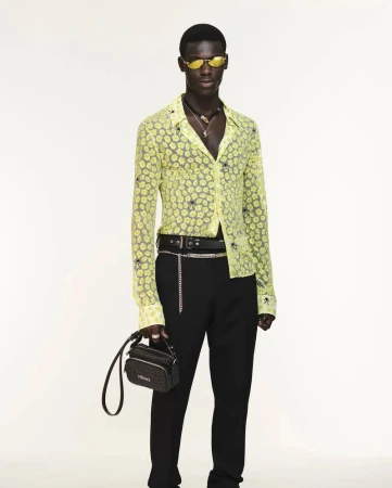 Full-body shot of a Black male model in black pants and a yellow patterned shirt, holding a bag, against a white backdrop in studio lighting.