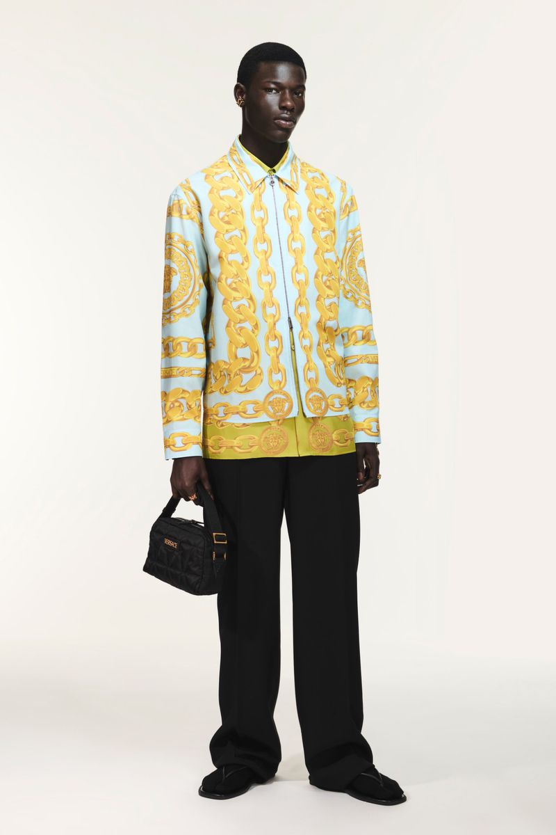 Full-length studio shot of a dark-skinned male model wearing a Versace shirt and black pants, holding a black Versace bag.