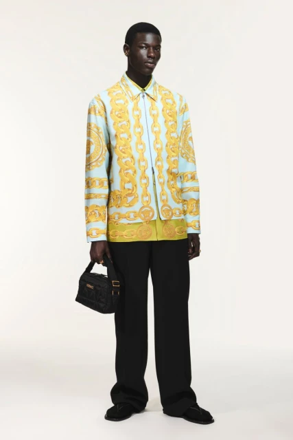 Full-length studio shot of a dark-skinned male model wearing a Versace shirt and black pants, holding a black Versace bag.
