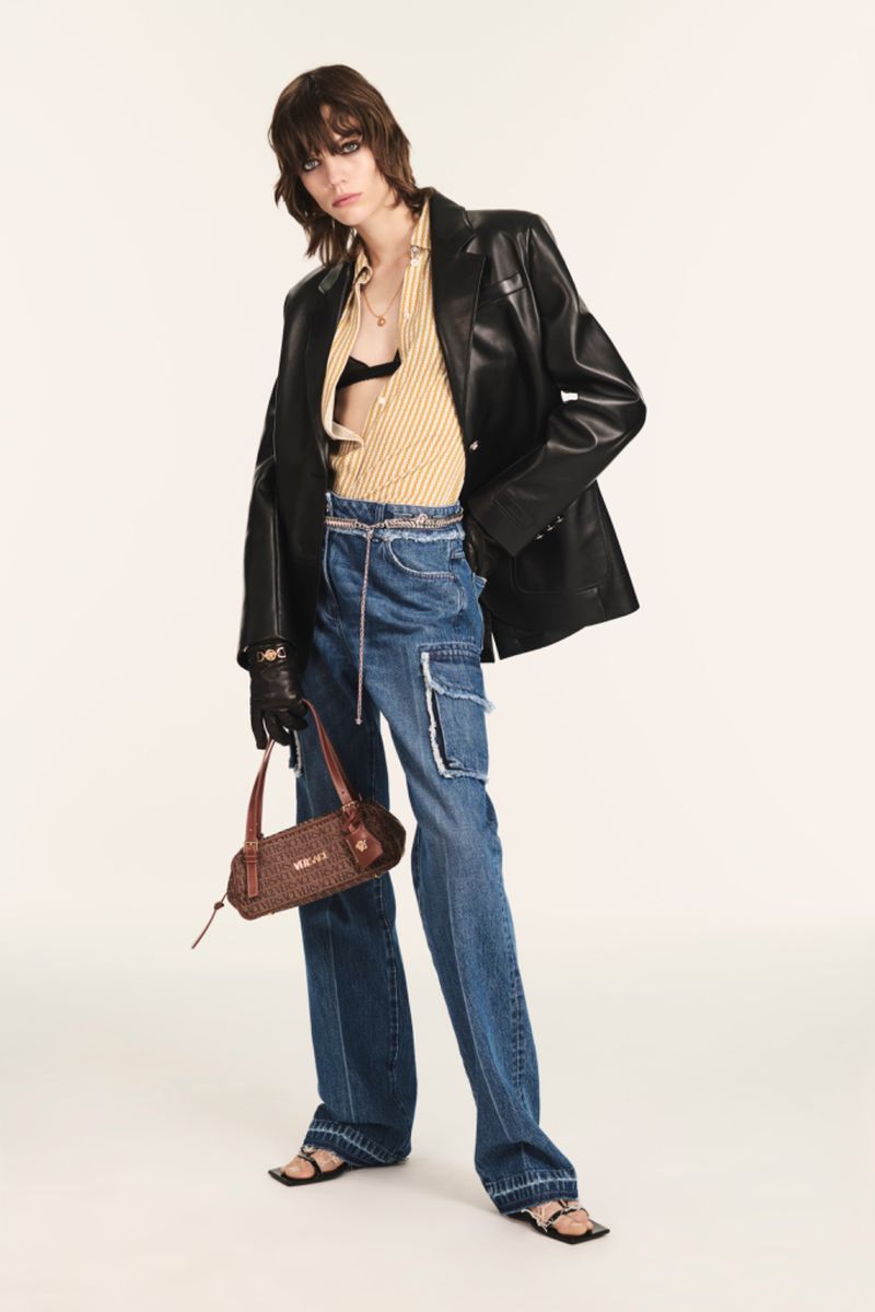 A woman poses in baggy jeans and a leather blazer, holding a brown handbag against a neutral backdrop in a fashion studio shot.
