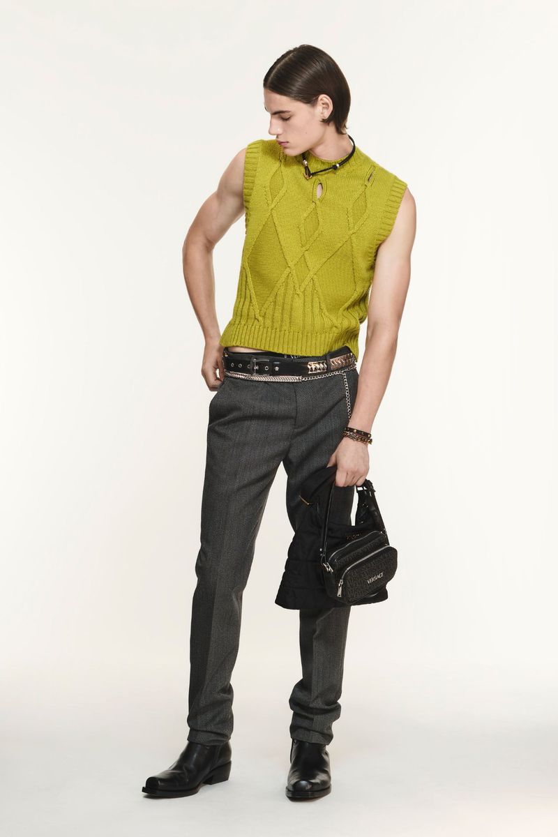 Male fashion model poses in a sleeveless knit vest, gray pants, and black boots, holding a small black bag on a white background.