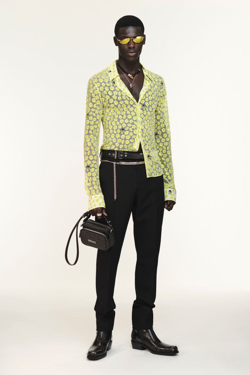 Black male model in a sheer yellow patterned shirt, black trousers, sunglasses, and leather boots holding a black handbag on white seamless.