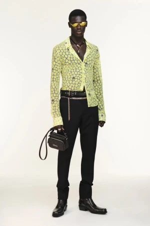 Black male model in a sheer yellow patterned shirt, black trousers, sunglasses, and leather boots holding a black handbag on white seamless.