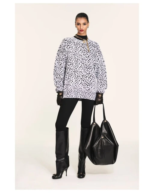 A woman models a Dalmatian-spotted sweatshirt with black leggings, high-heeled boots, a large black handbag, and gold chain necklace.