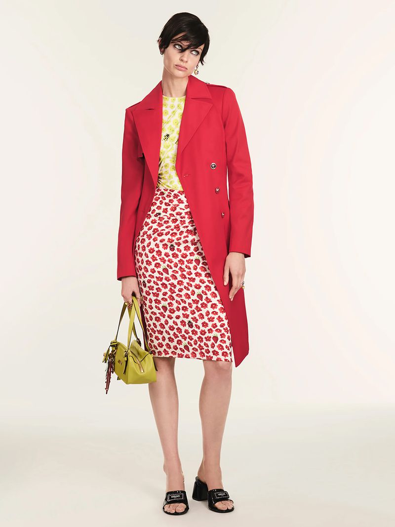 A stylish woman models a red trench coat over a yellow top and red floral skirt, accessorized with a green bag and black heeled mules.