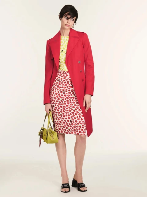 A stylish woman models a red trench coat over a yellow top and red floral skirt, accessorized with a green bag and black heeled mules.
