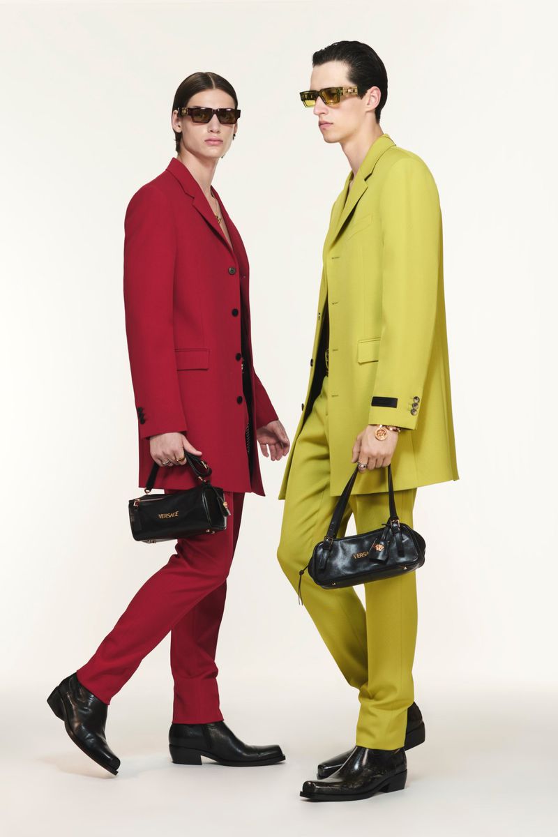 Two men in red and lime-green suits walk side-by-side in a studio wearing sunglasses and carrying black bags in a fashion editorial.
