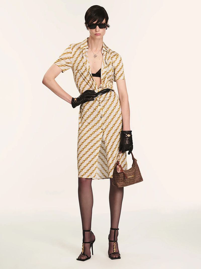 A fair-skinned woman poses in a chain-pattern dress, gloves, and heels, holding a handbag against a plain background in a fashion shot.