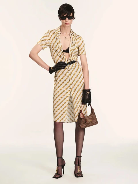 A fair-skinned woman poses in a chain-pattern dress, gloves, and heels, holding a handbag against a plain background in a fashion shot.