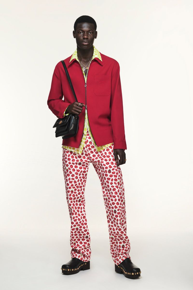 A male model stands in a red jacket, flower-print pants, black bag, and clogs against a white backdrop in a full-length studio shot.