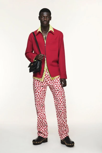 A male model stands in a red jacket, flower-print pants, black bag, and clogs against a white backdrop in a full-length studio shot.