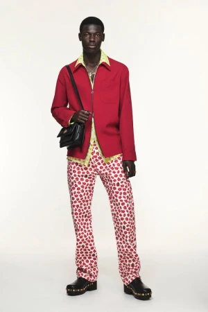 A male model stands in a red jacket, flower-print pants, black bag, and clogs against a white backdrop in a full-length studio shot.