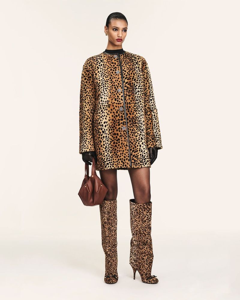 Fashion model in a leopard print coat dress and matching knee-high boots holding a brown handbag against a white backdrop.
