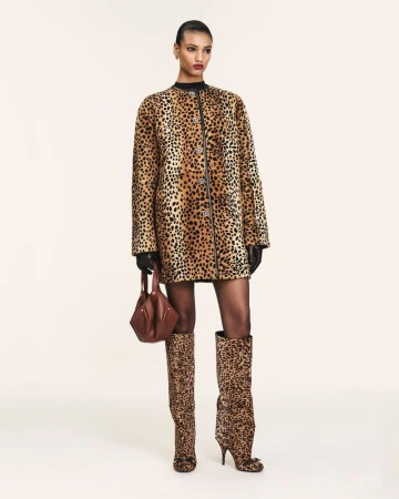 Fashion model in a leopard print coat dress and matching knee-high boots holding a brown handbag against a white backdrop.