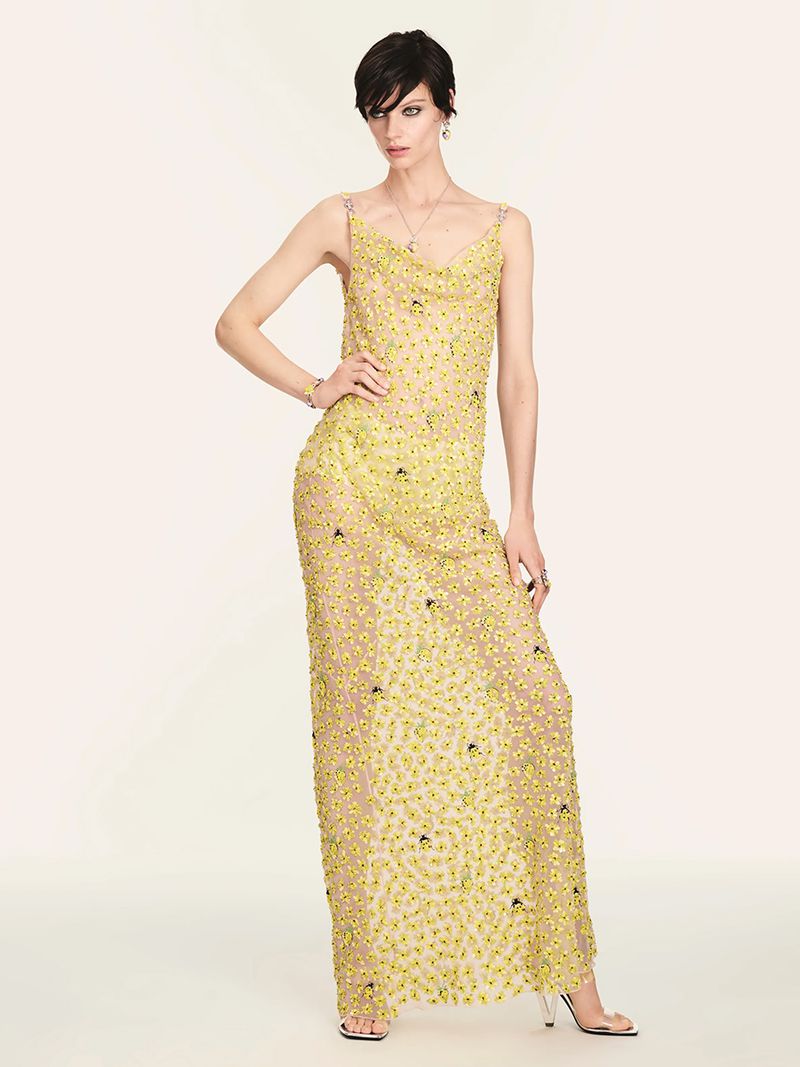 Fashion model in a sheer yellow beaded gown poses in a studio. The full-length shot emphasizes the gown's delicate details and design.
