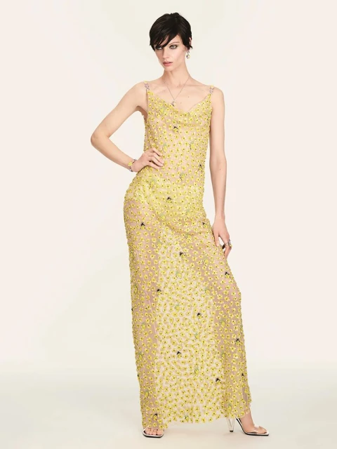 Fashion model in a sheer yellow beaded gown poses in a studio. The full-length shot emphasizes the gown's delicate details and design.