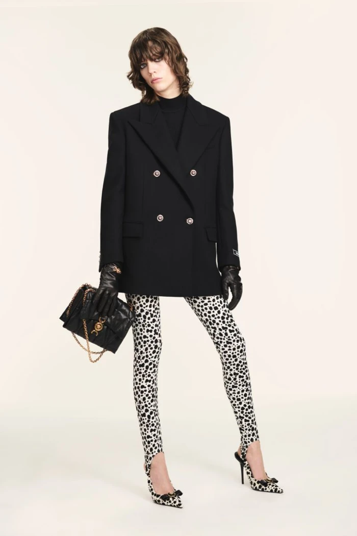 Model wearing a black double-breasted blazer and animal-print leggings holding a black handbag against a plain white seamless backdrop