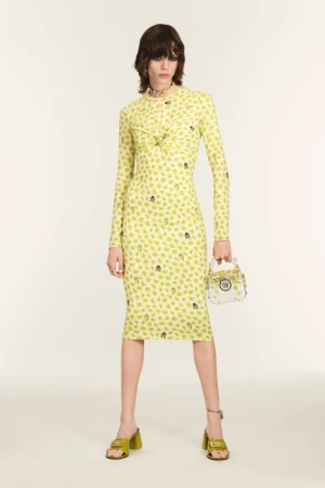 Model wearing a pale yellow floral long-sleeve midi dress with lime green mules and a white handbag against a light studio backdrop.