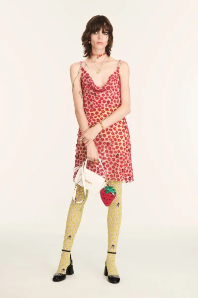 Model wearing a red floral mini dress, patterned yellow tights, and black heels holding a white handbag with a strawberry charm.